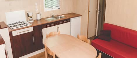 Et kompakt tekøkken med gasblus, vask og lille køleskab. Spisebord med stole og en rød sofa findes i opholdsstuen.