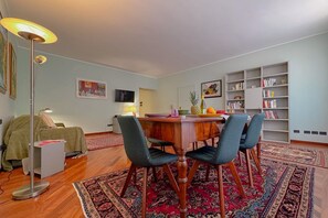 A dining area with a wooden table, two chairs, a floor lamp, a bookshelf, and a television.