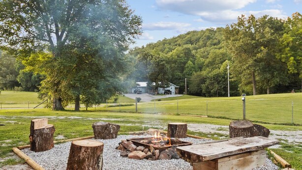 A fire pit with logs and a stone bench in a grassy area with trees and a house in the background. - Direct Access to Brimstone Trails & River. Hot Tubs. Fire Pits. Pets (Huntsville)