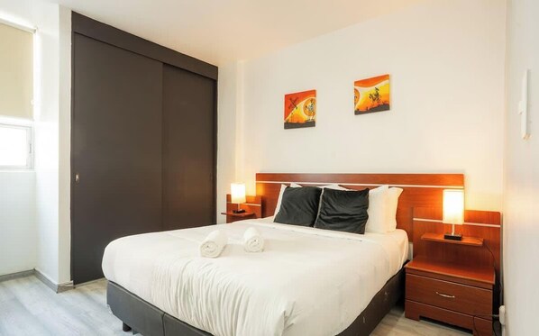 Una habitación de hotel con una cama grande, dos mesitas de noche con lámparas, una puerta corredera oscura y dos cuadros colgados en la pared.