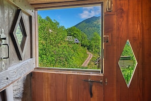 Une porte en bois ornée d’une fenêtre triangulaire, laissant entrevoir un paysage verdoyant et luxuriant, avec une route sinueuse et une maison.