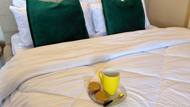 A neatly made bed with white bedding, two green pillows, a yellow mug, and two cookies on a clear glass plate.