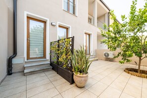 Un bâtiment doté d’un patio carrelé, d’une plante en pot, d’un portail en métal noir et d’un petit arbre planté dans un bac en bois.