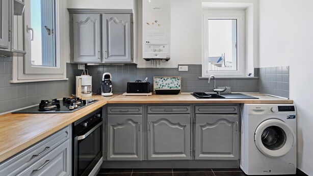 A kitchen with grey cabinets, a built-in washing machine, a coffee maker, and a window. - Holidays in Perros-Guirec – Relaxation, Sea, and Discovery! (Perros-Guirec)