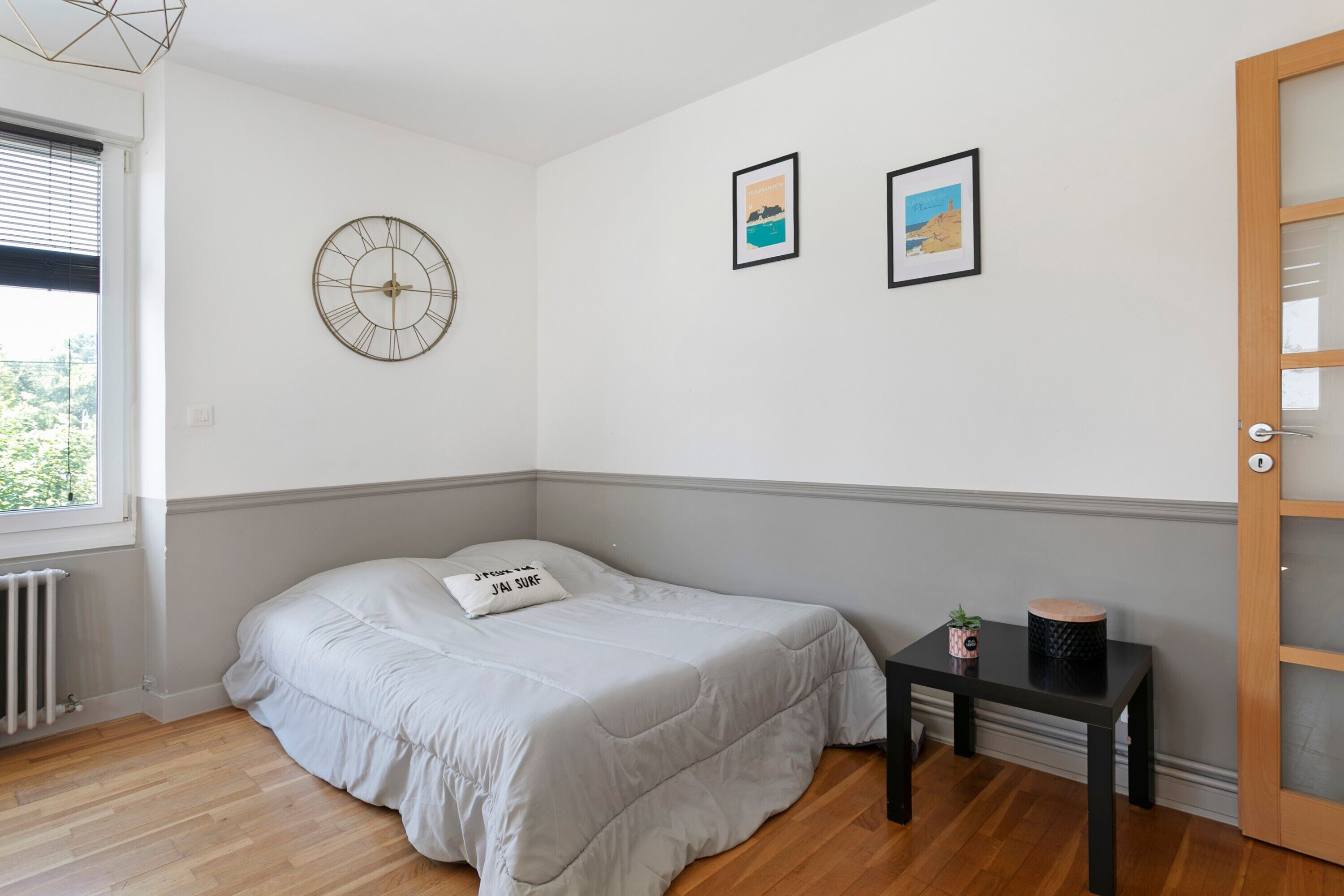 A bedroom with a bed, a bedside table with a plant, a clock, and two framed pictures on the wall.