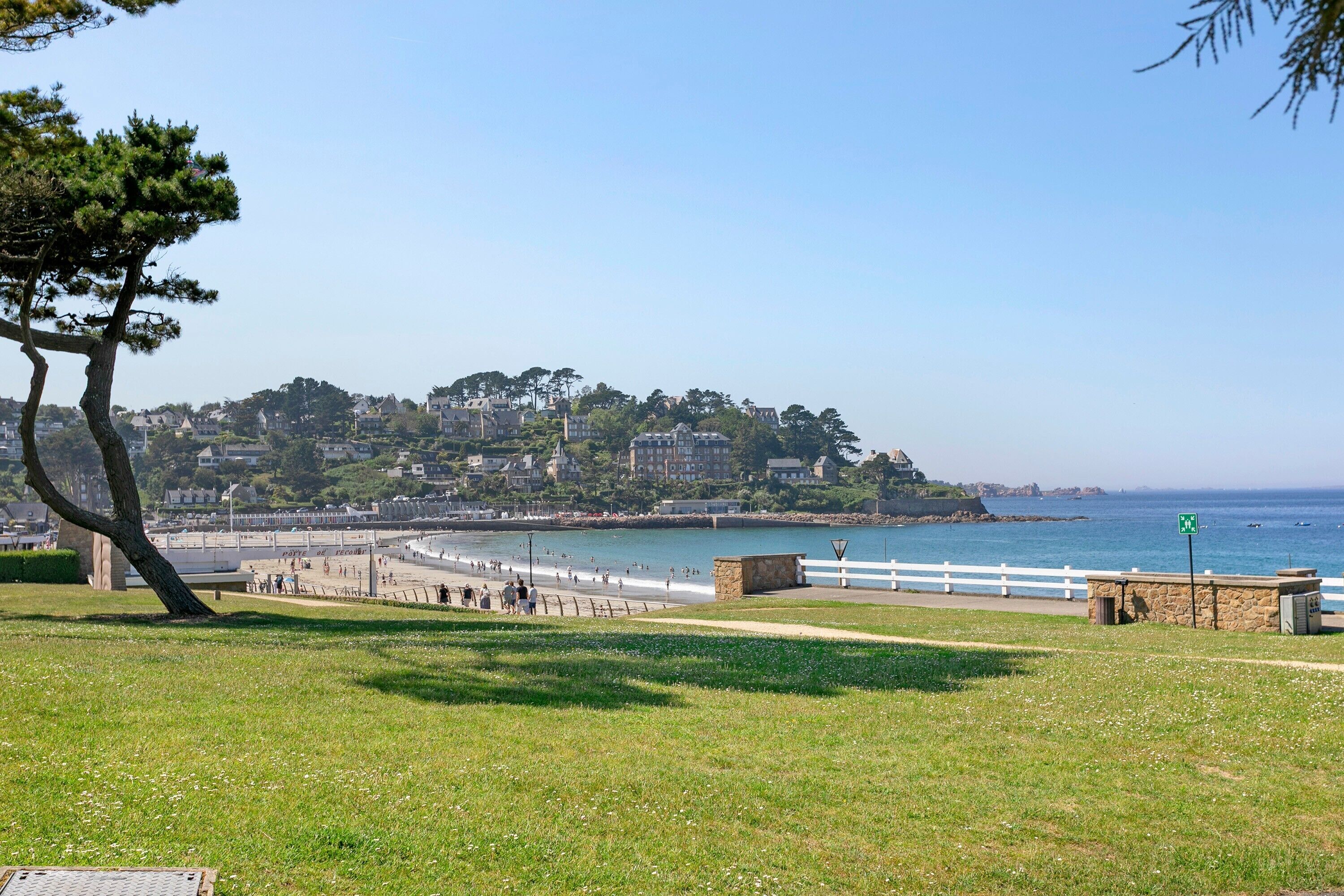 A coastal park with a grassy area, a stone bench, and a white fence along the beach.