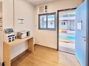 A room with a wooden desk, a microwave, and a digital device. There is a door leading to a brightly colored area outside.
