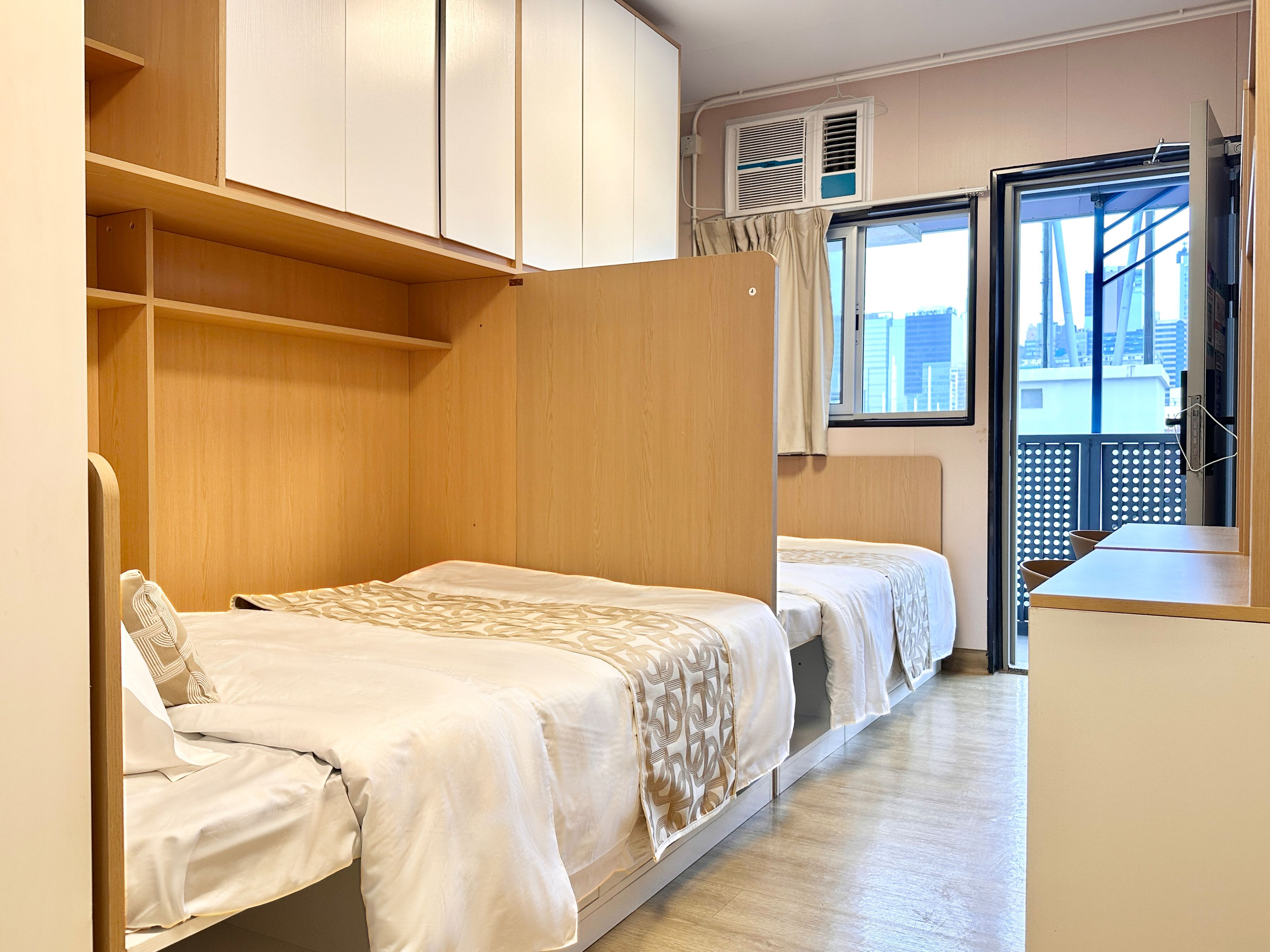 A compact room with two beds, a wardrobe, and a window with a view of buildings.