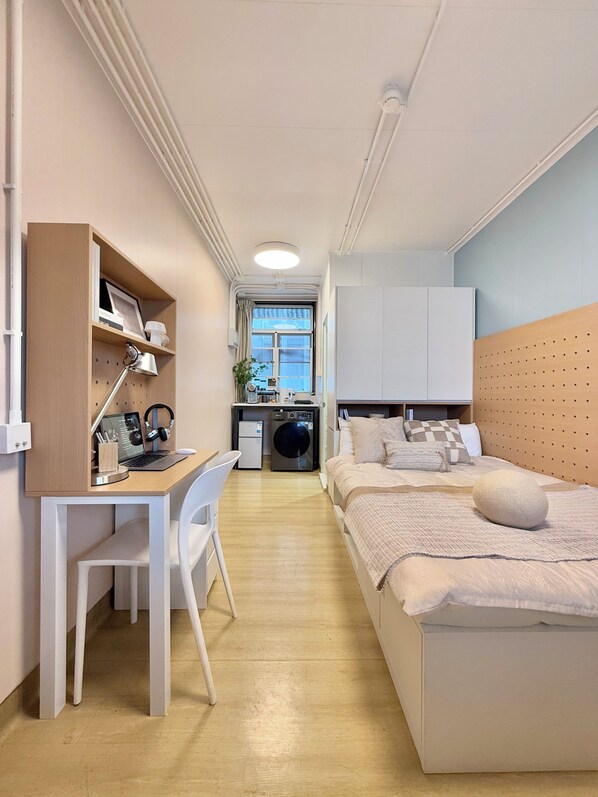 A compact room with a bed, a desk with a chair, a washing machine, and a small kitchenette.