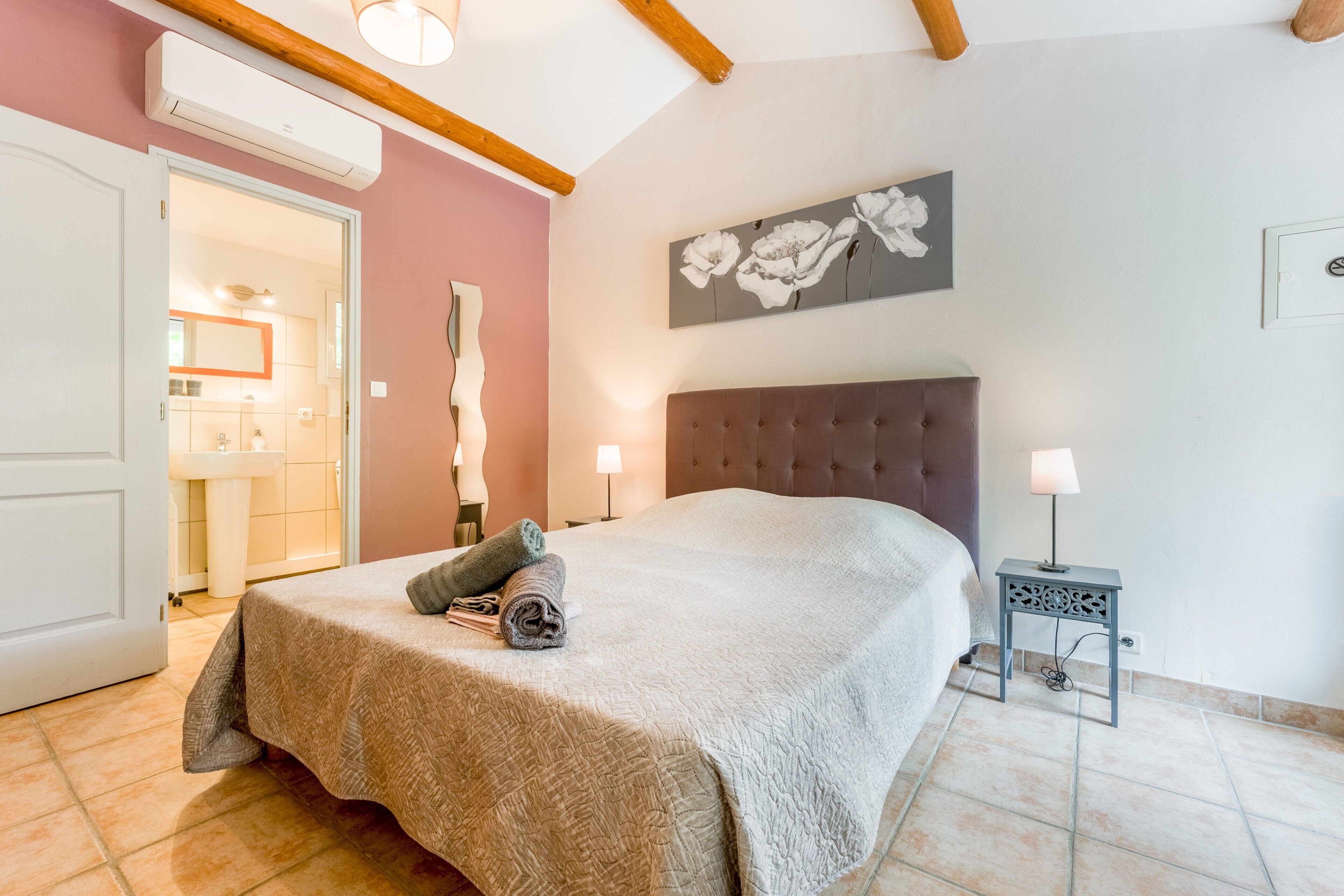 A bedroom with a bed, a bedside table with a lamp, a wall-mounted air conditioner, and a bathroom visible through an open door.