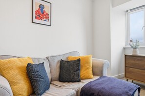 A living room with a sofa, cushions, a framed artwork, a window, and a wooden dresser.