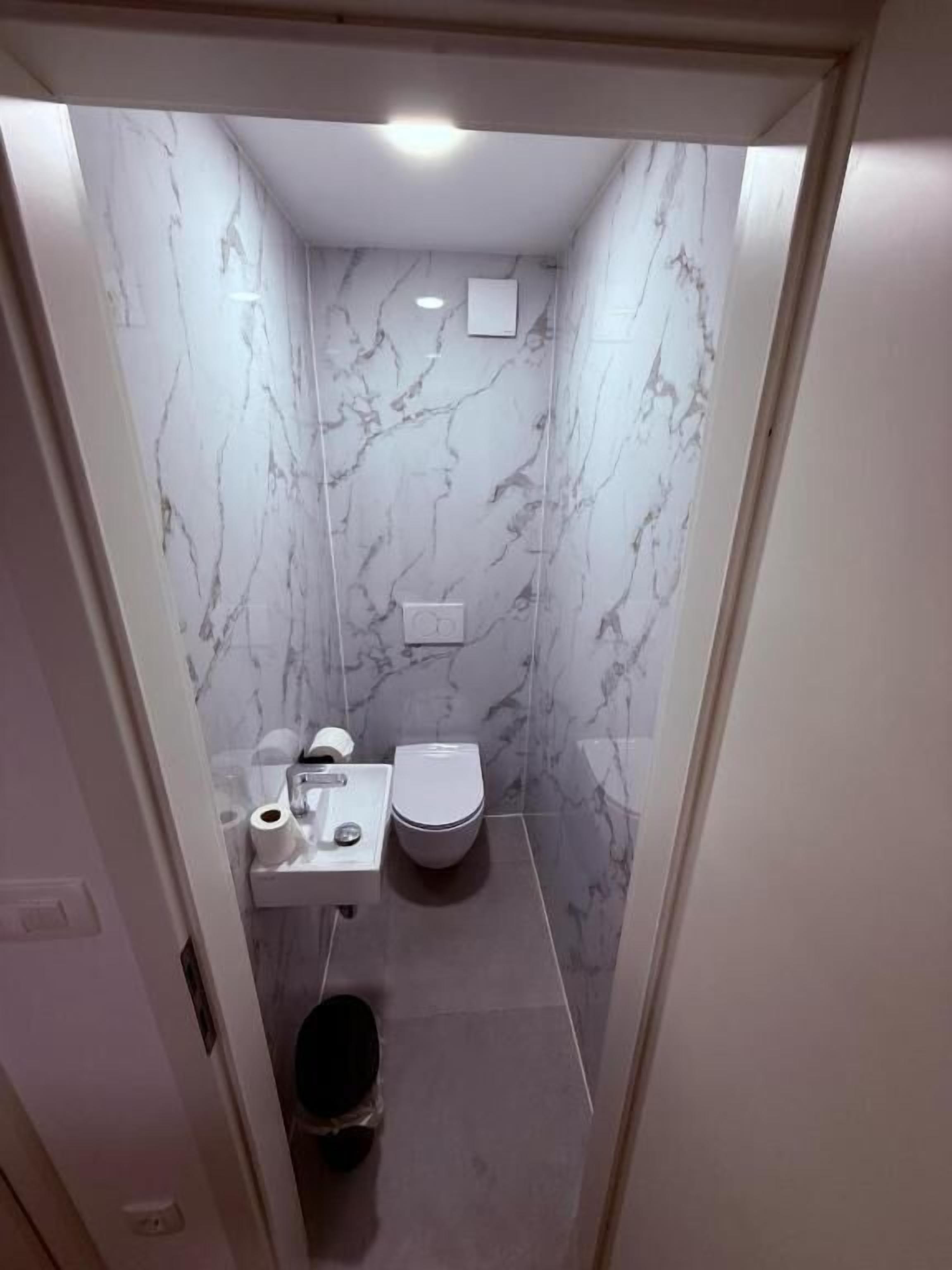 A narrow bathroom with marble walls, a toilet, a sink, and a trash bin.