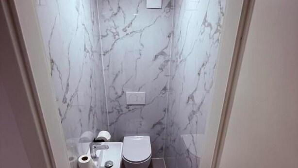 A narrow bathroom with marble walls, a toilet, a sink, and a trash bin. - PJ Resort - Pool & Mountain View (Cerklje na Gorenjskem)
