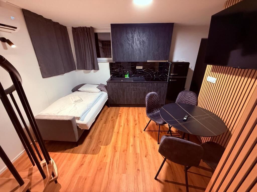 A compact room with a bed, a small kitchenette, a dining table with chairs, and a wooden partition.