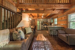 Ein gemütliches Blockhaushaus-Interieur mit einer Holztreppe, einem Wohnzimmer mit Sofa und Sesseln, einer Küchenzeile inklusive Kühlschrank und Herd sowie einem großen Fenster.