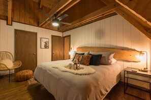 Une chambre avec un grand lit, un ventilateur de plafond, un plafond en bois, une table de chevet et une chaise.