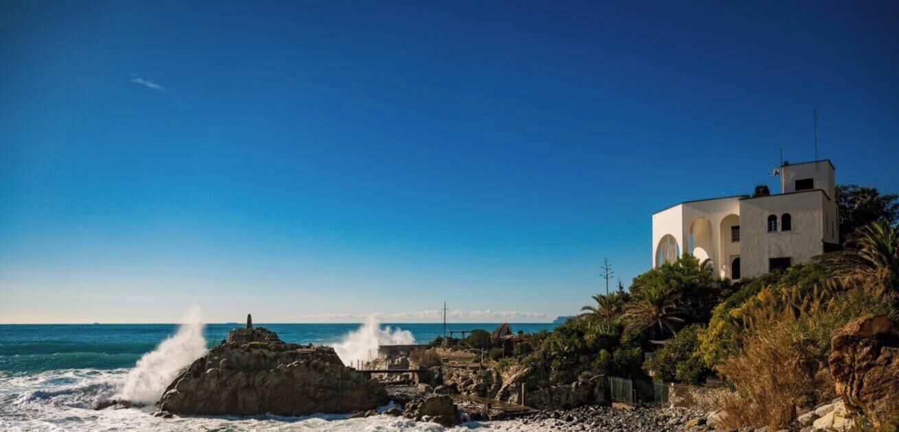 Un edificio costiero con finestre ad arco, un litorale roccioso e onde che si infrangono sulle rocce.