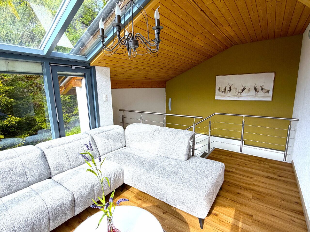 Ein modernes Wohnzimmer mit einem Ecksofa, einer Chandelierleuchte, einer Holzbalkendecke, einem Balkon mit Metallgeländer und einem Bild von Hirschen an der Wand.