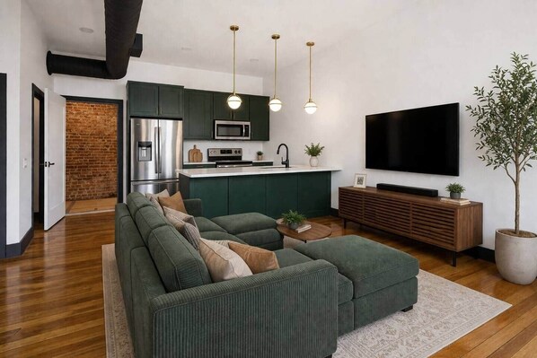 A modern living room with a sectional sofa, a flat-screen TV, a kitchen area with a microwave, and a large potted plant.