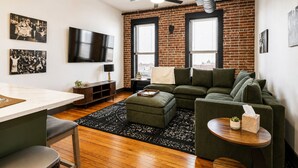 Ein modernes Wohnzimmer mit Backsteinwand, einem grünen Sofa, einem Holztisch und einem an der Wand montierten Fernseher.