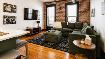 A modern living room with a brick wall, a green sofa, a wooden coffee table, and a television mounted on the wall.