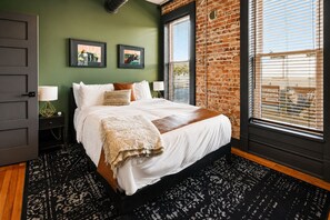 A bedroom with a brick wall, a large bed, two framed pictures, and a window with blinds. - Reasonable Doubt --- Vibrant, unforgettable loft in Downtown Port Clinton (Port Clinton)