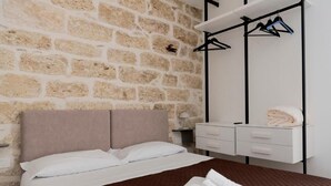 Una habitación de hotel con pared de piedra, una cama con colcha marrón, una mesita de noche blanca con lámpara y un armario con perchas negras.