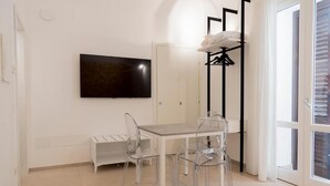 Una habitación con una mesa blanca, sillas transparentes, un televisor montado, un perchero y un ventanal de vidrio corredero.