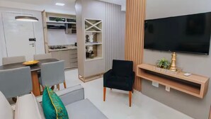 Een moderne woonkamer met een bank, een aan de muur bevestigde televisie, een eethoek met een tafel en stoelen, en een keuken op de achtergrond.