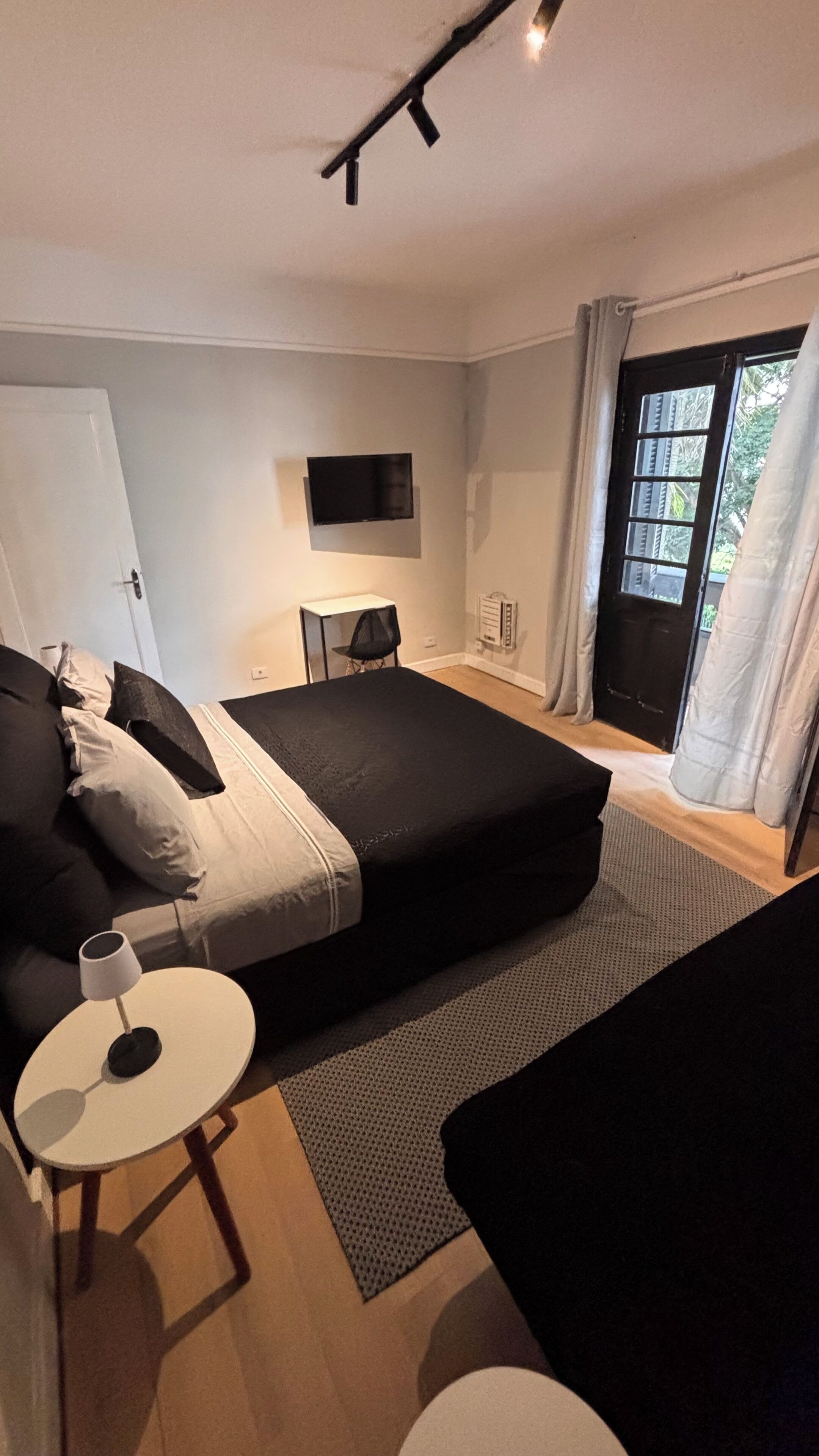 Un dormitorio con cama, una mesita, una silla, un televisor y un ventanal corredero.