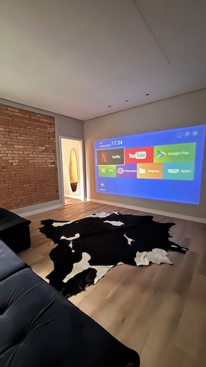 Una sala de estar con una pantalla grande que muestra una interfaz multimedia, un sofá de cuero, un tapete de piel de vaca y una pared de ladrillo.