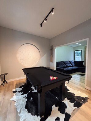 Et billardbord med kugler, en ruskindst æppe og en moderne sofa i en stue med lyse vægge og en loftlampe.