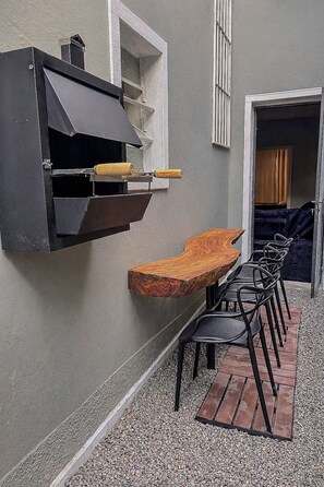 En grill, en træbænk og en række stole i et udendørs opholdsområde.