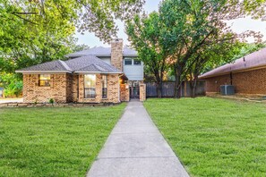 A two-story house with a brick exterior and a stone chimney, a front yard with a concrete pathway, and a tree-lined driveway. - Cozy Frisco Getaway - Fireplace + Relaxing Backyard (Frisco)