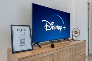Ein Fernseher mit dem Disney+-Logo, ein gerahmtes Schild mit dem WLAN-Symbol und ein kleiner Dekorationsventilator auf einer Holzschrank.