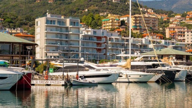 Un port de plaisance avec des bateaux amarrés, un immeuble à plusieurs étages et un paysage vallonné en arrière-plan.