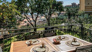 Une table en bois, dressée pour quatre personnes, avec des sets de table, un bol et des couverts, sur un balcon donnant sur une vue panoramique de la ville.