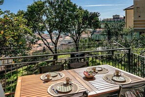 Ein Holztisch für vier Personen, gedeckt mit Platzsets, einer Schüssel und Besteck, auf einem Balkon mit Blick auf eine Stadtlandschaft.