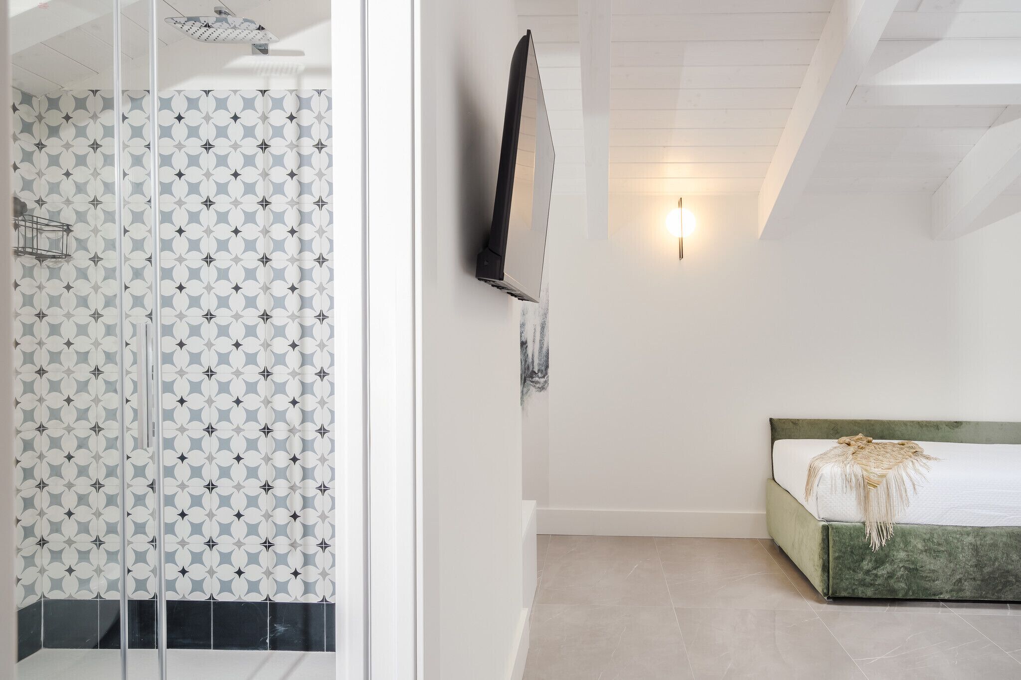 A bedroom with a bed, a TV, a wall-mounted light, and a patterned shower area.