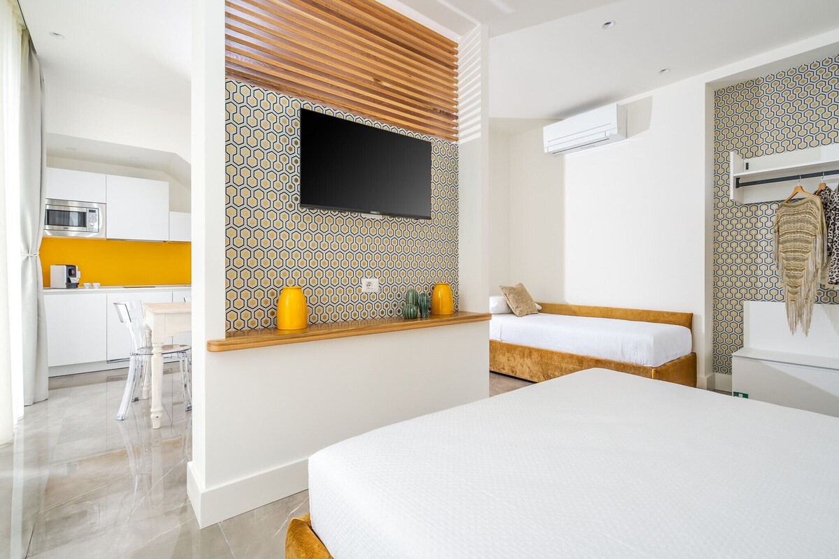 A modern hotel room with a bed, a small kitchenette, a TV, and patterned walls.