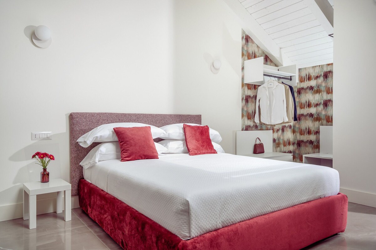 A modern bedroom with a bed, a nightstand, a vase of flowers, and a wall-mounted mirror.