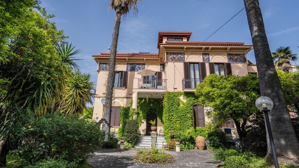 Un edificio de dos pisos con techo de tejas, un balcón y un jardín con diversas plantas y una palmera.