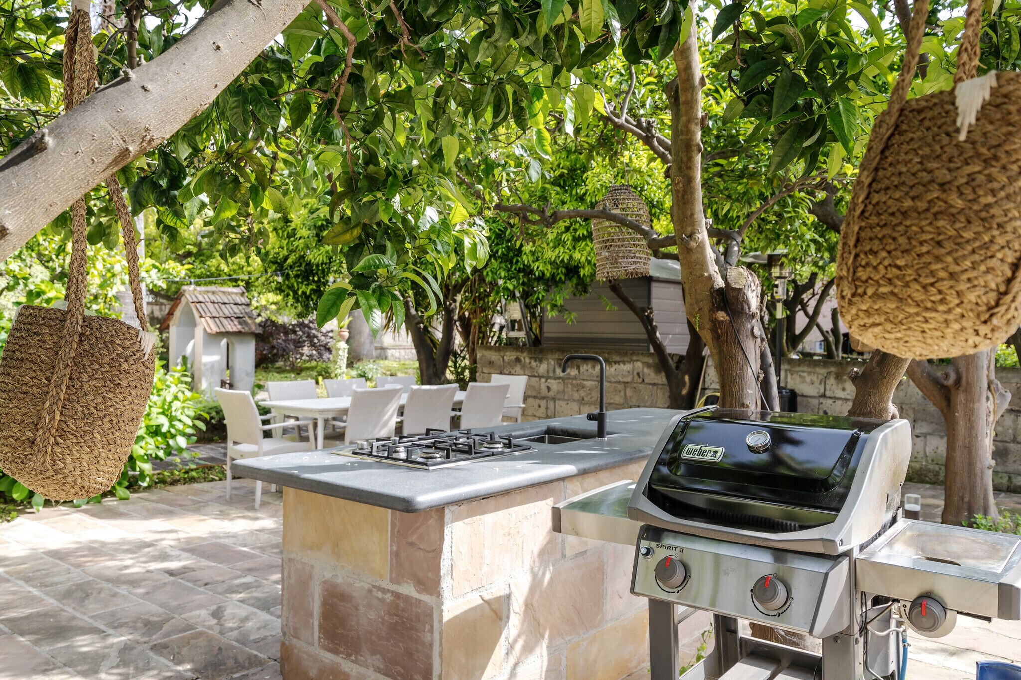 Cocina exterior con una parrilla Weber, encimera de piedra, cestas colgantes y zona de patio con sillas y mesas.