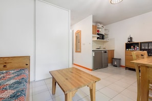 A compact living space with a bed, a wooden table, a kitchen area with appliances, and a small trash bin.