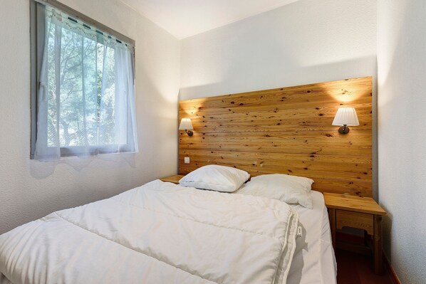 A bedroom with a wooden headboard, two bedside lamps, a window with sheer curtains, and a bed with white bedding.