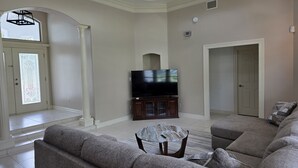 Ein Wohnzimmer mit einem Ecksofa, einem Flachbildfernseher auf einem Holzkasten, einem Glastisch und einem Deckenventilator.