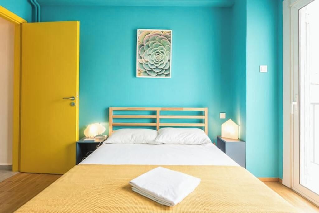 A room with a bed, a yellow door, a teal wall, and a painting.