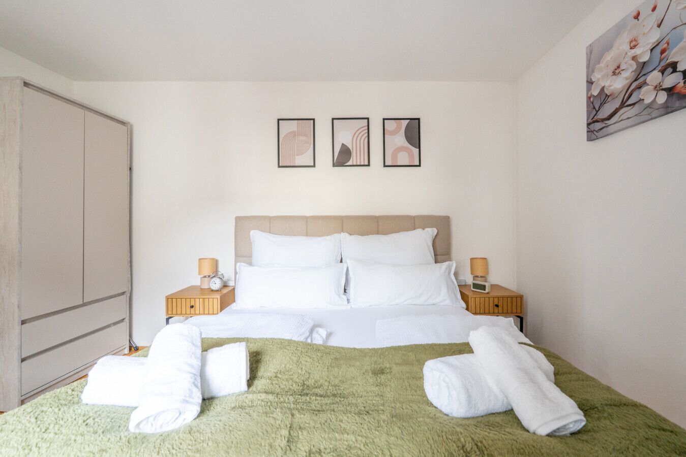 A hotel room with a bed, two bedside tables with lamps, a large wardrobe, and three framed pictures on the wall.