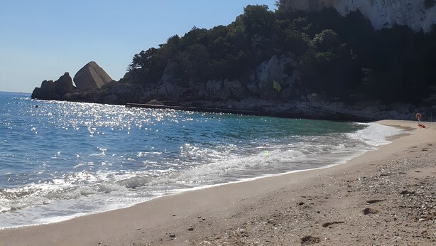A beach with clear skies, a large rock formation, and calm sea.