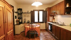 A kitchen with wooden cabinets, a dining table with chairs, a window with blinds, and a hanging light fixture.